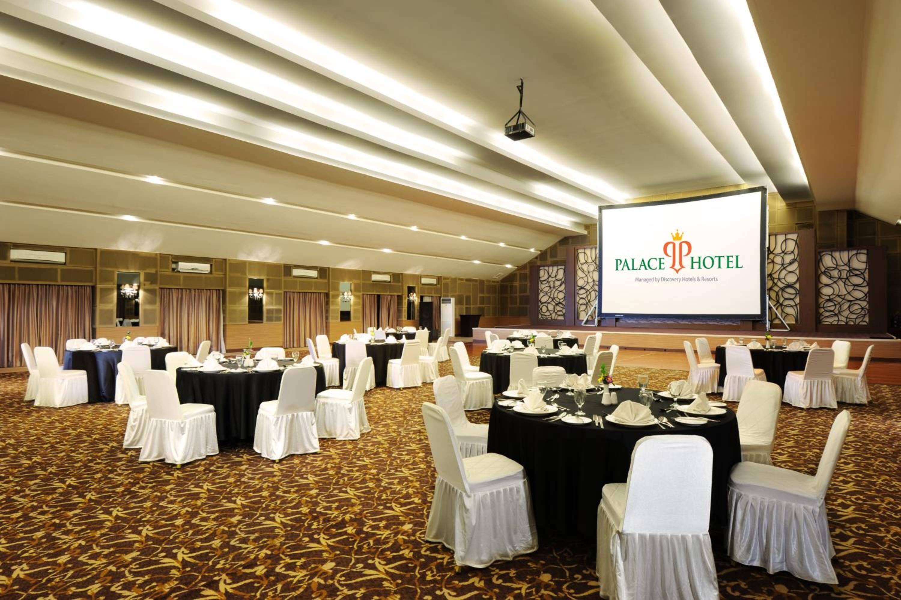 Un salón de banquetes con mesas redondas y sillas blancas, una pantalla grande mostrando la marca 'Palace Hotel', y un suelo alfombrado.