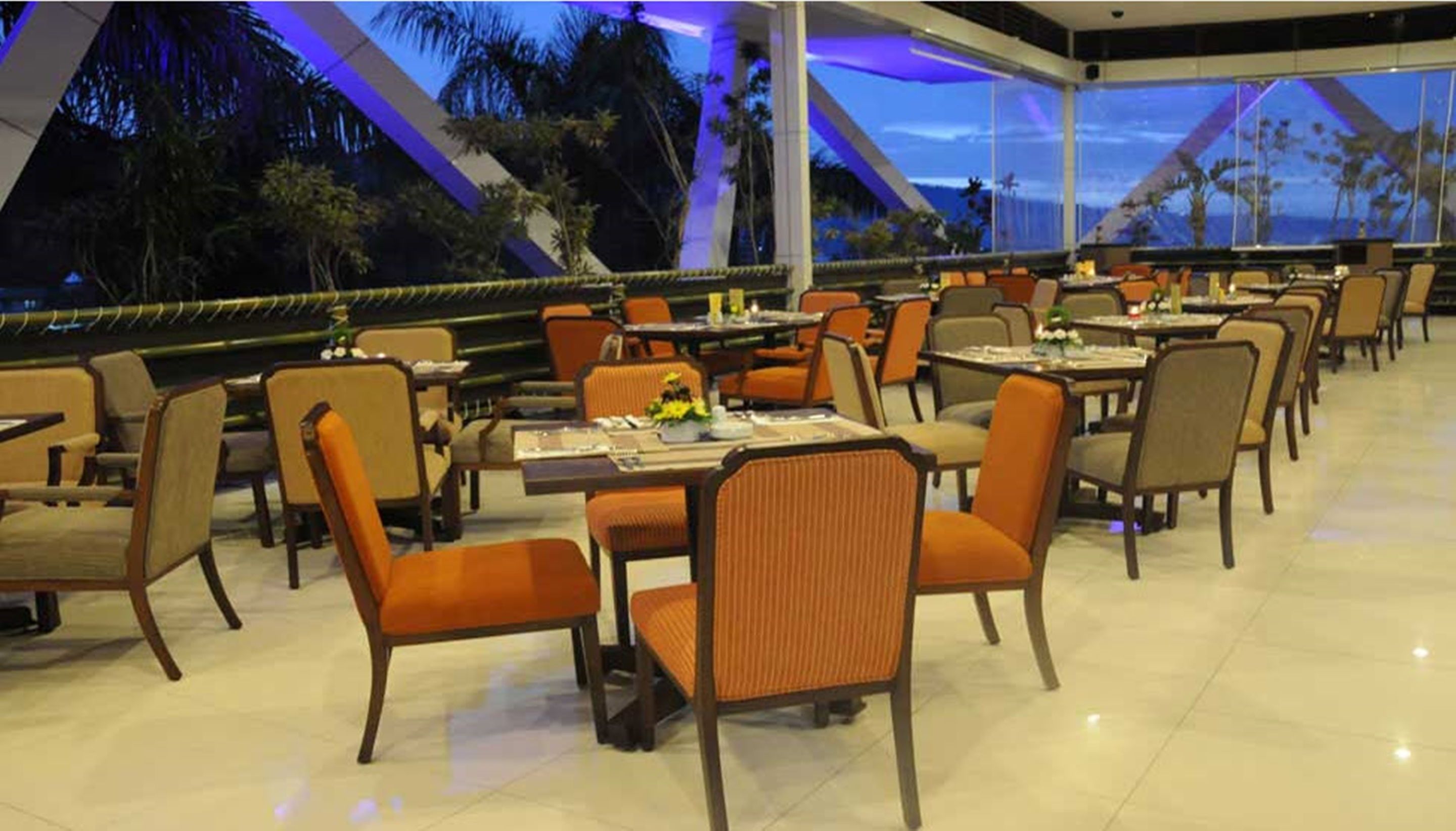 A restaurant with multiple tables and chairs, a modern architectural design, and a view of palm trees.