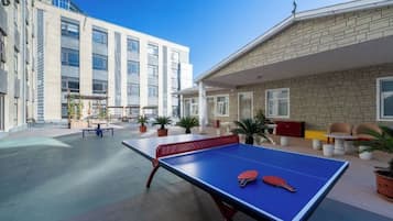Mesa de ping pong al aire libre con una red, plantas en macetas y un edificio con grandes ventanales al fondo.