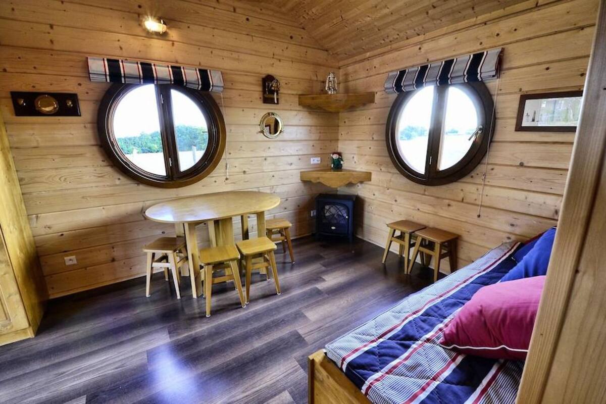Interior acogedor de una cabaña con una mesa redonda para comer, dos sillas de madera, un taburete pequeño, una cama con una colcha a rayas y dos ventanas grandes con contraventanas.