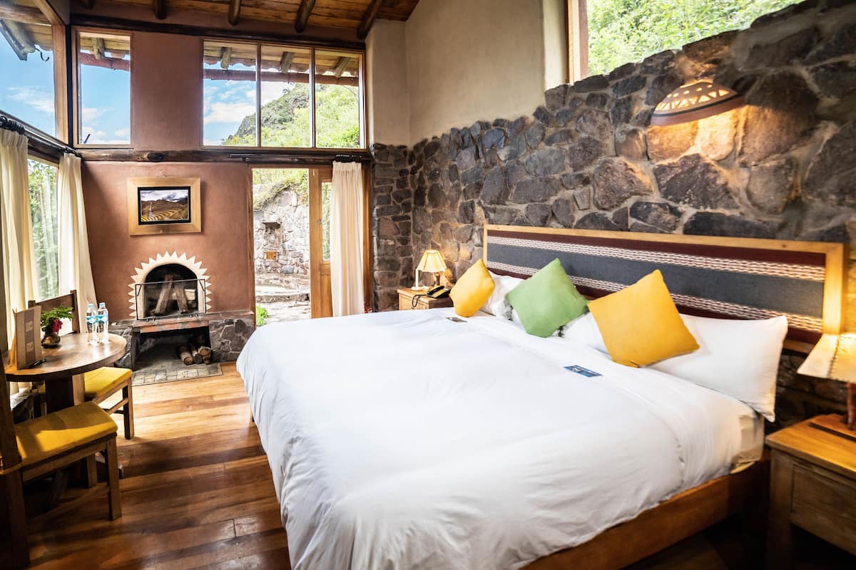 A bedroom with a stone wall, a fireplace, a bed with pillows, a wooden desk, and a window with curtains.