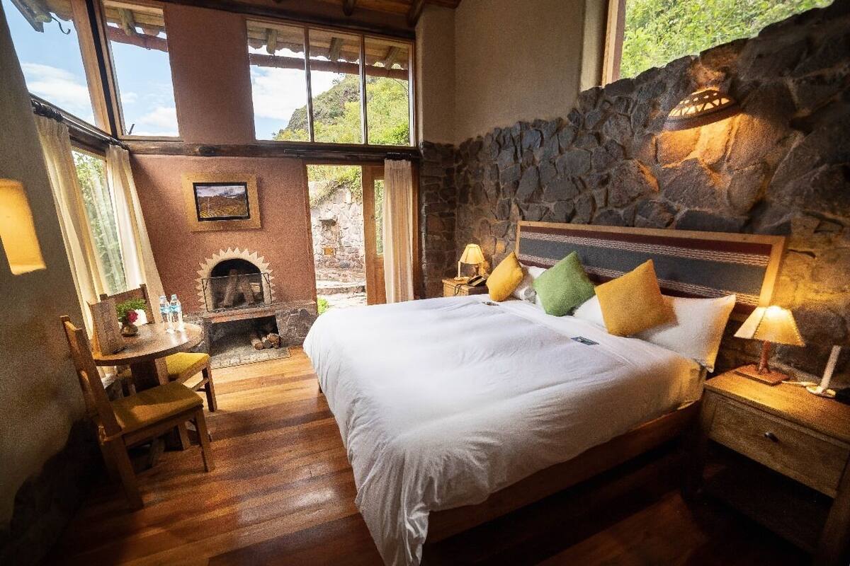 A bedroom with a stone wall, a fireplace, a bed with pillows, a nightstand, a table with a chair, and a window with a view of greenery.