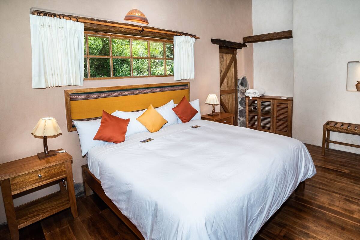 A neatly made bed with a wooden headboard, a nightstand with a lamp, a bench, and a window with curtains.
