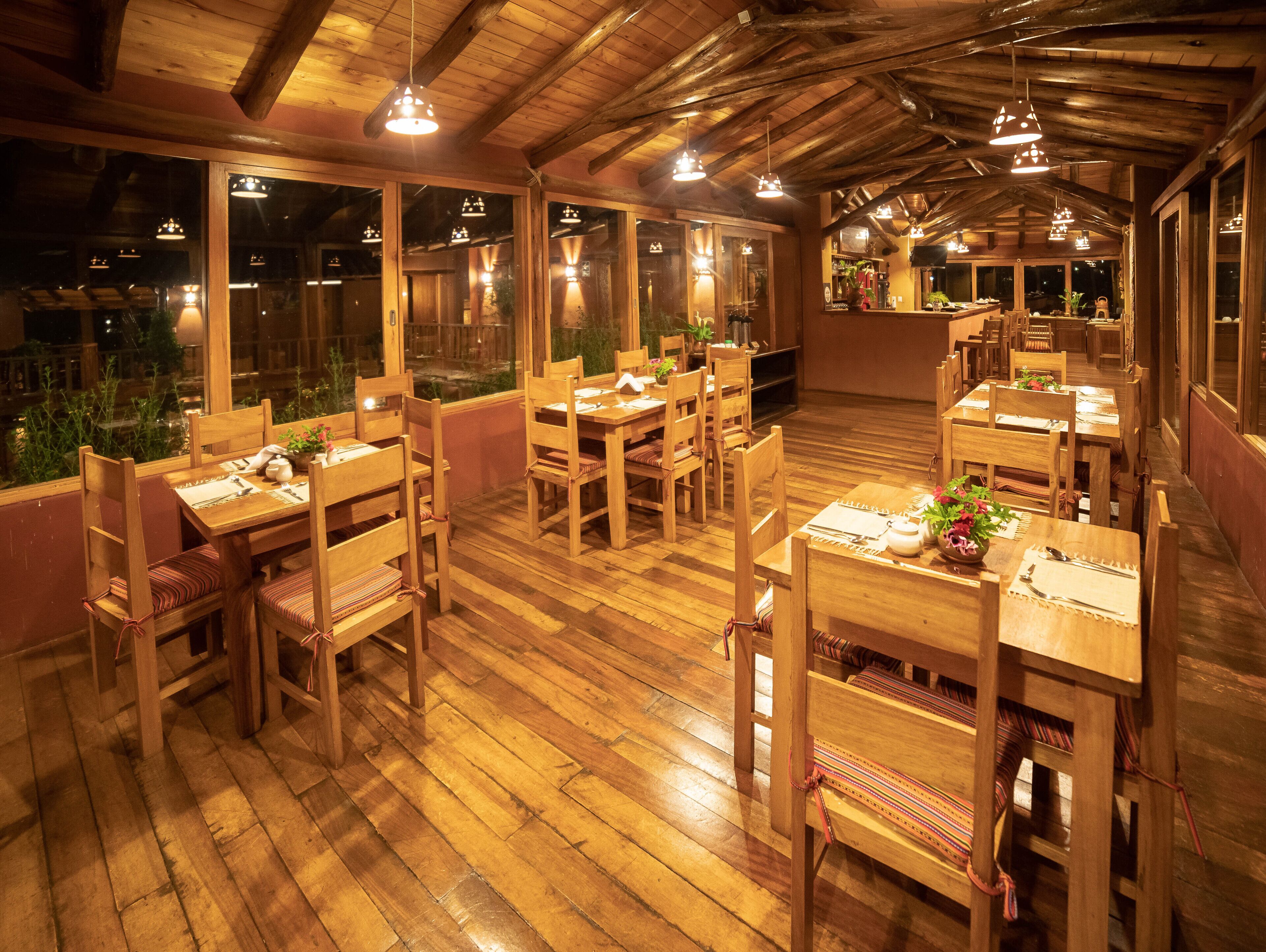 Un restaurante con mobiliario de madera y ventanas amplias. Las mesas están preparadas con platos y cubiertos.