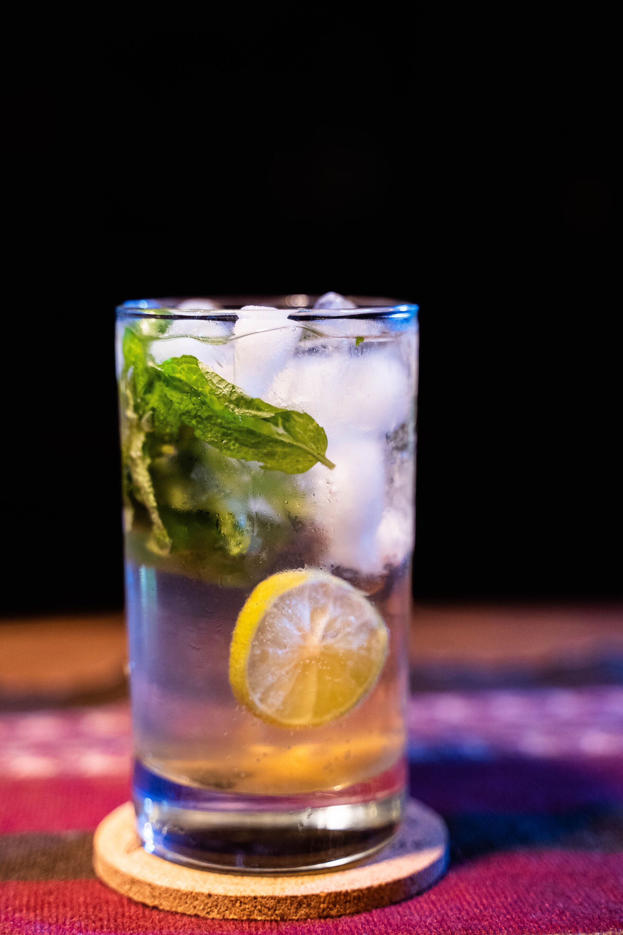 A glass of iced beverage with mint leaves and a lemon slice on a coaster.
