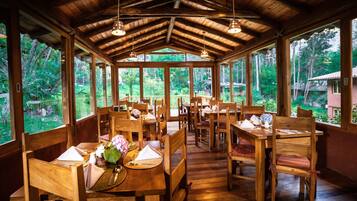 Un comedor de madera con mesas dispuestas para cuatro, ventanas amplias y vistas naturales al exterior.