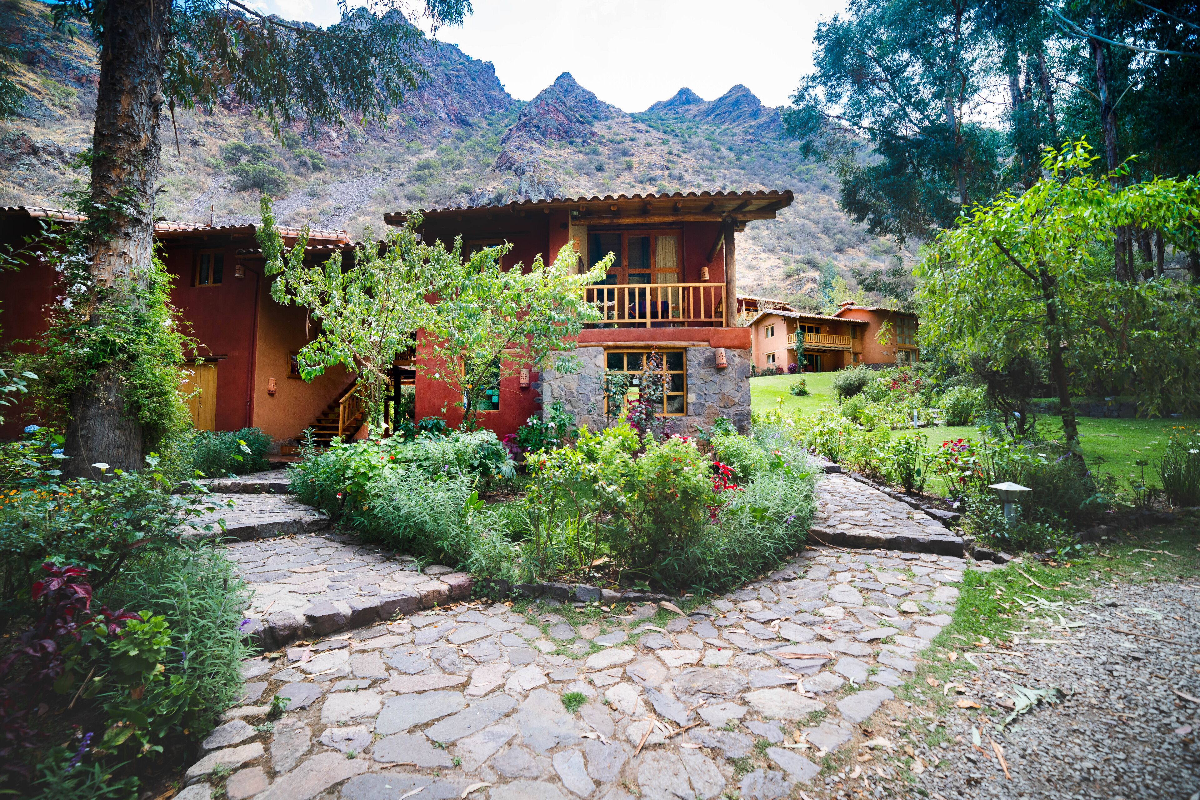 A rustic lodge with a stone pathway leading to a wooden cabin, surrounded by lush gardens and mountains in the background.