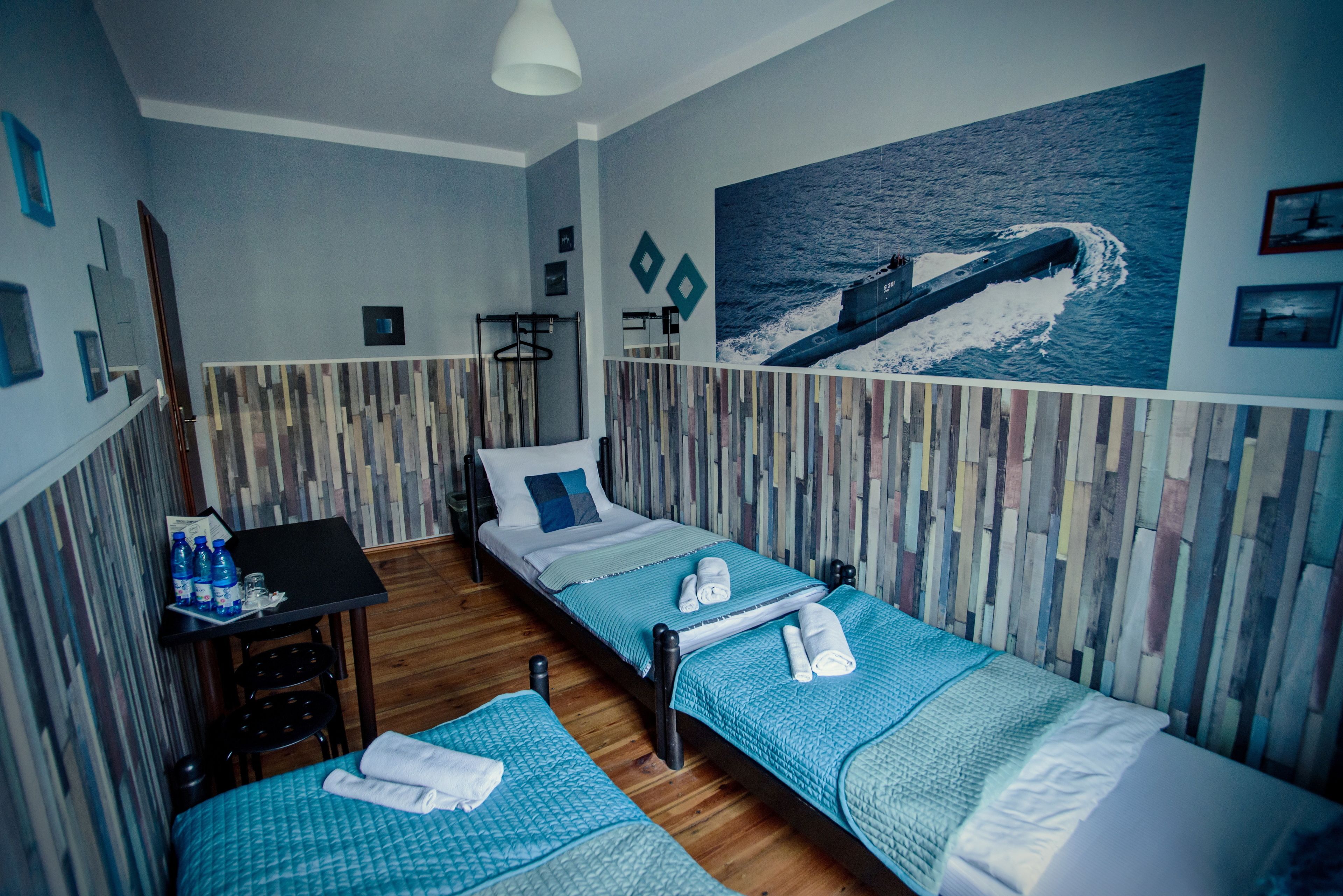 Une chambre d’hôtel avec deux lits simples, une petite table avec une chaise, un bureau avec une chaise et une grande fresque murale sur le thème de l’océan.
