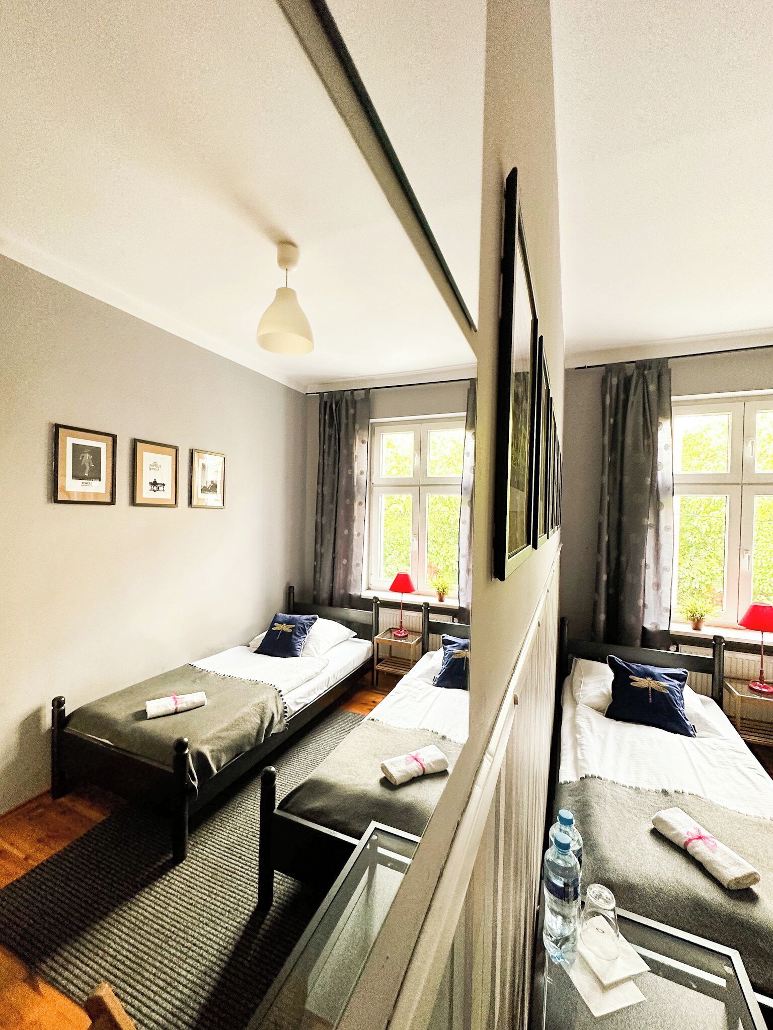 Une chambre avec deux lits simples, une petite table avec une lampe rouge, une fenêtre avec des rideaux et trois tableaux encadrés accrochés au mur.
