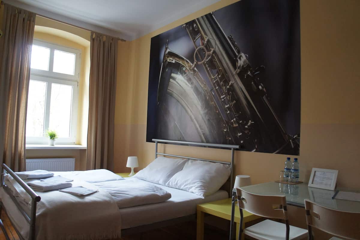 Hotellihuone, jossa on sänky, työpöytä, tuoli, ikkuna verhoilla ja suuri juliste seinällä.