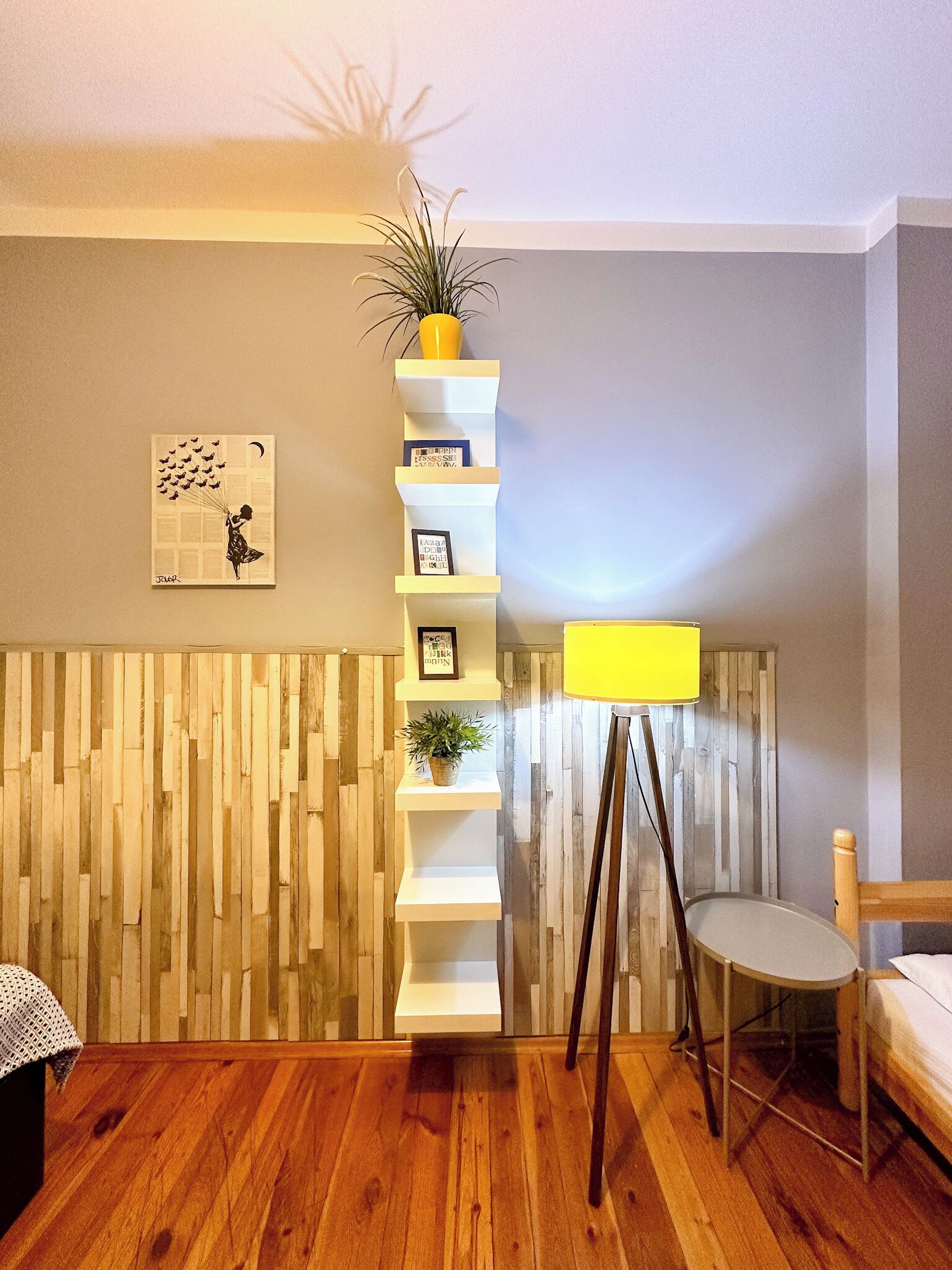 A room with a wooden floor, a white wall, a vertical shelf unit, a small table, and a chair.