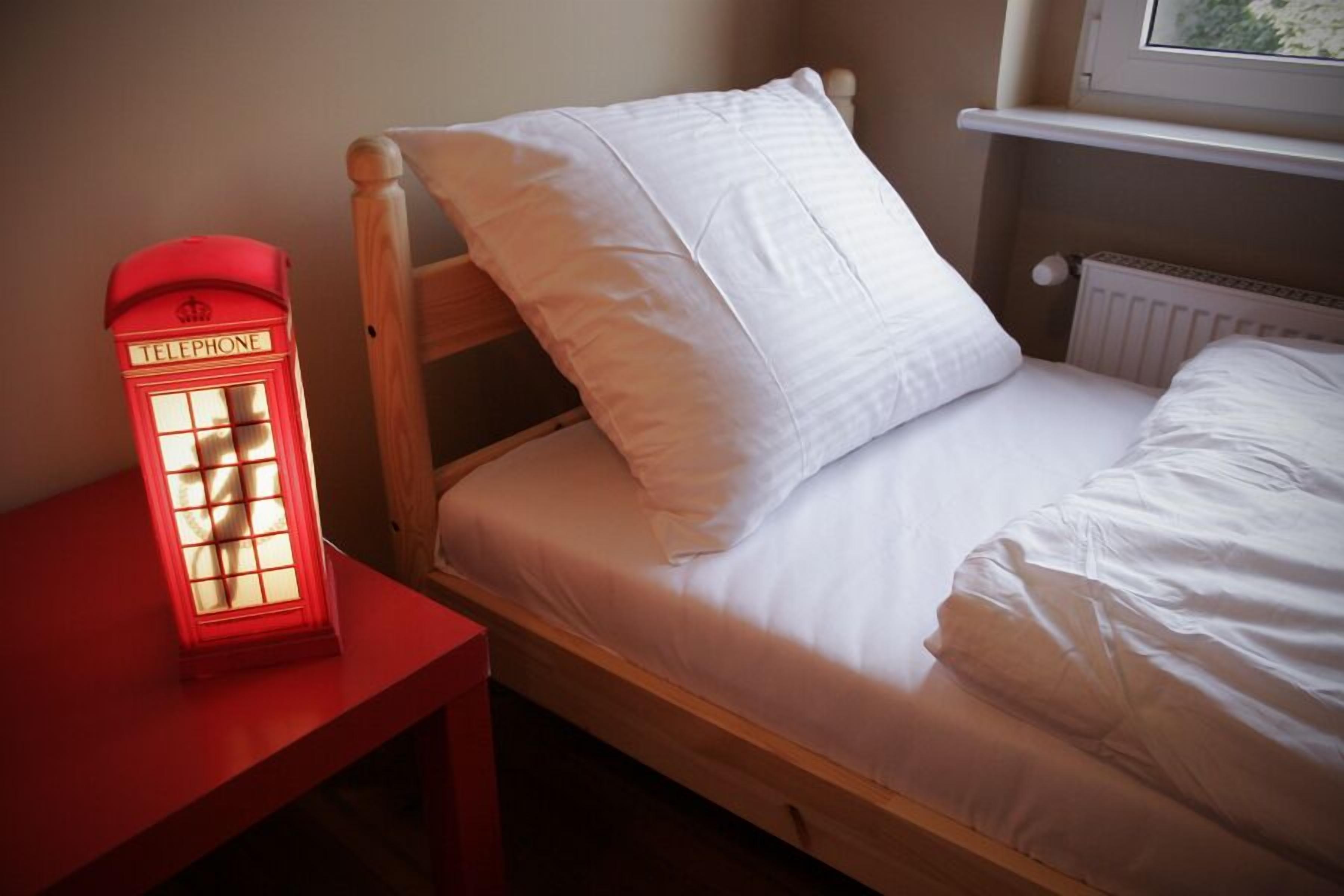 Un lit simple avec une literie blanche, une tête de lit en bois et une lampe de chevet de style cabine téléphonique rouge.