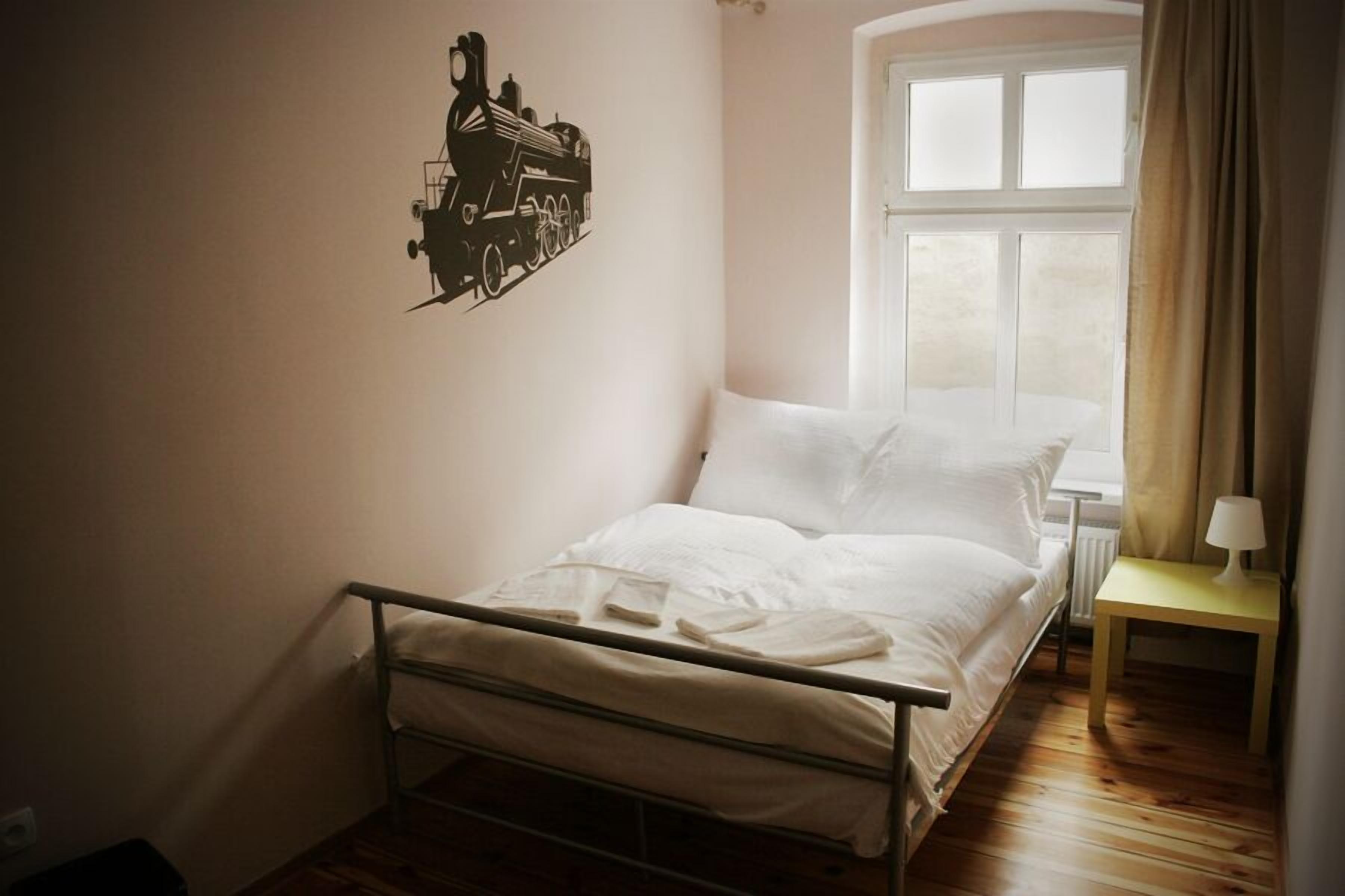 A single bed with a metal frame, a bedside table with a lamp, a window with curtains, and a wall decal of a train.