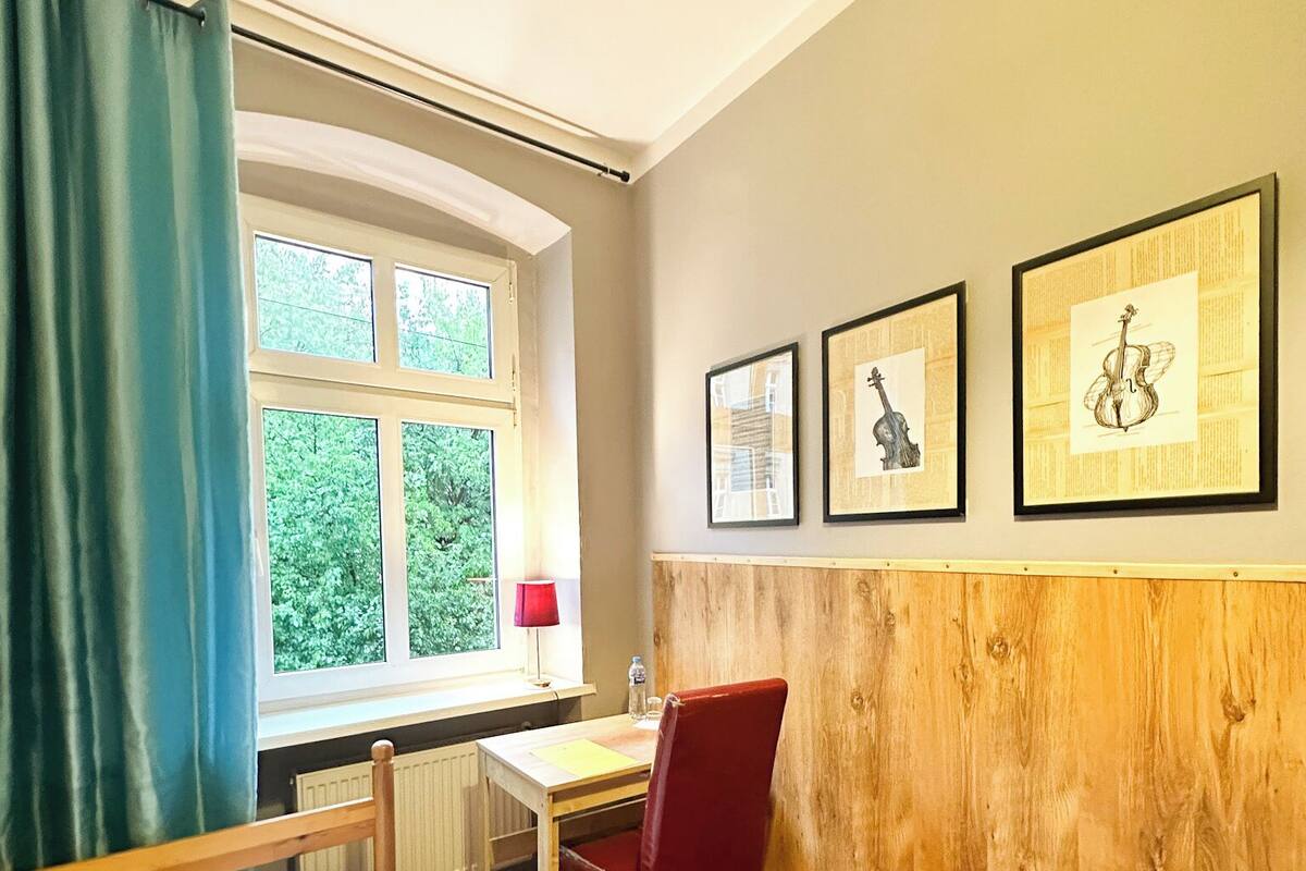 Hotellihuone, jossa on sänky, työpöytä, tuoli, ikkuna verhoilla ja seinällä olevia kehystettyjä tauluja.