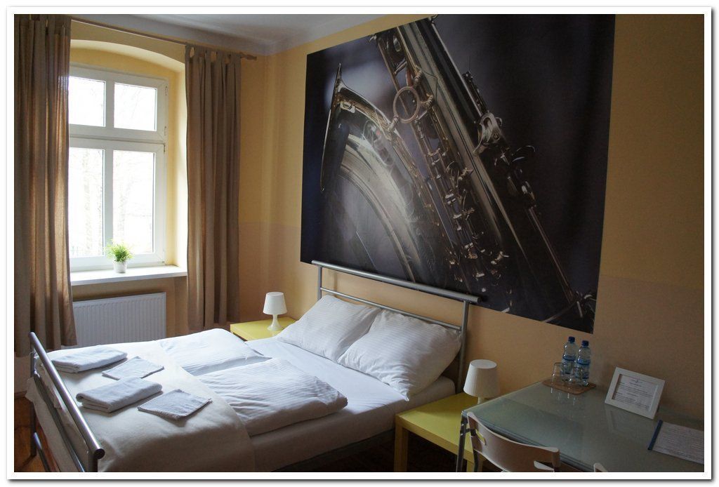 A hotel room with a bed, a window with curtains, a wall-mounted picture, a bedside table with a lamp, and a table with water bottles.