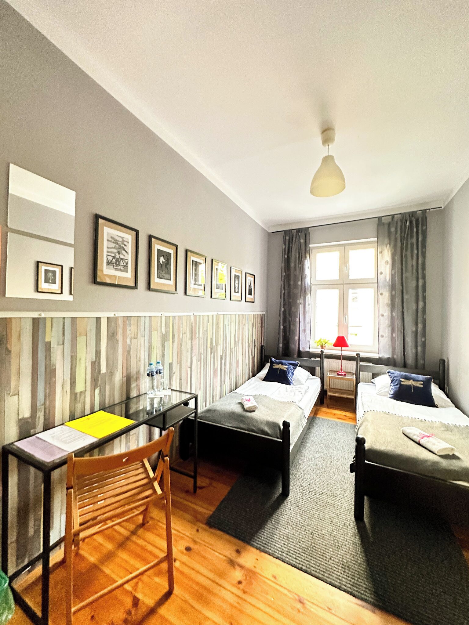 A hotel room with two single beds, a desk with a chair, a wooden floor, a window with curtains, and framed pictures on the wall.