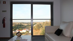 Une pièce avec un canapé blanc, une petite table sur laquelle se trouvent un vase et des livres, et une grande fenêtre donnant sur un paysage rural.