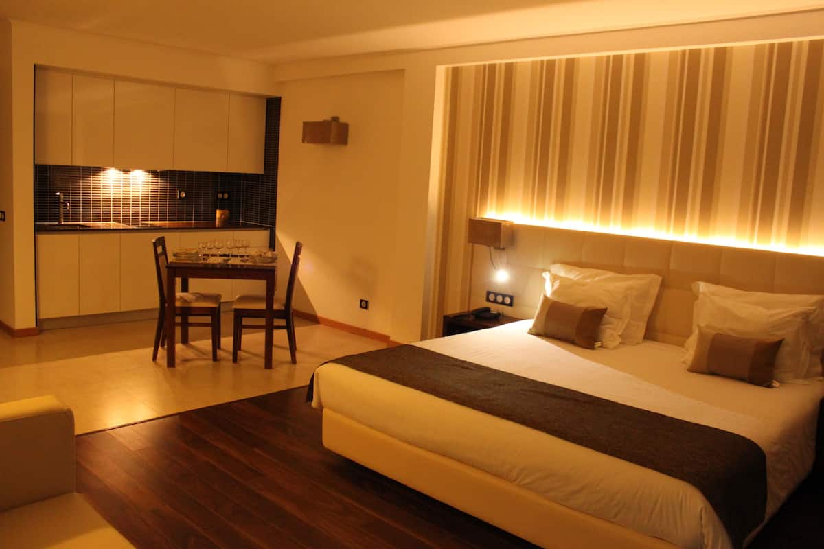 Een hotelkamer met een bed, een eettafel met stoelen, een kitchenette en een groot raam met gordijnen.