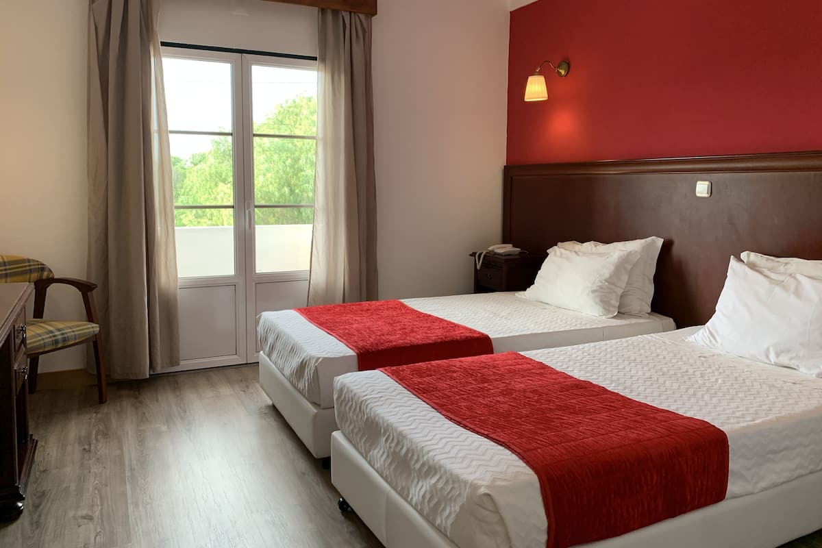 Hotelkamer met twee bedden, een houten hoofdbord, een rode accentmuur, een raam met gordijnen, een nachtkastje met een lamp en een stoel.