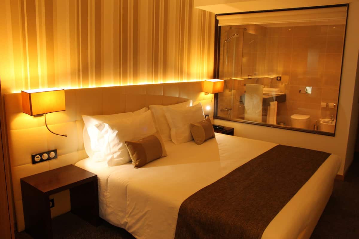 Hotelkamer met een groot bed, bedlampen, een nachtkastje en uitzicht op de badkamer door een glazen wand.