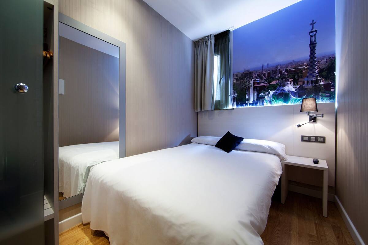 Habitación de hotel con una cama grande, una cama más pequeña, una mesita de noche, una lámpara y vistas a un paisaje urbano a través de una ventana.