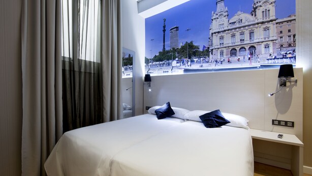 A hotel room with a bed, a nightstand, a lamp, and a view of a historic building. - Hostal Bcn Ramblas (Barcelona)