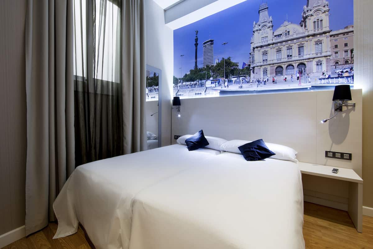 Habitación de hotel con cama, mesita de noche, lámpara y vista a un edificio histórico.