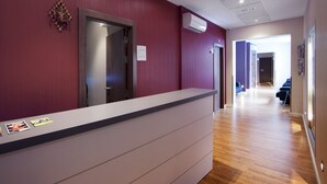 A reception area with a wooden reception desk, a red wall, and a hallway leading to a room with a bed. - Hostal Bcn Ramblas (Barcelona)