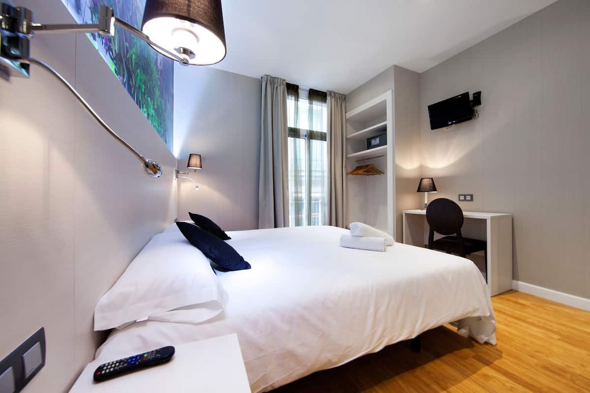Habitación de hotel con una cama grande, un escritorio, una silla, un televisor en la pared y una ventana con cortinas.