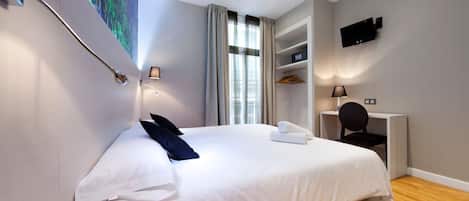 Habitación de hotel con una cama grande, un escritorio, una silla, un televisor en la pared y una ventana con cortinas.