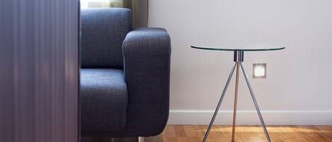 A modern room with a dark blue armchair, a glass-top table with metal legs, and a wooden floor.