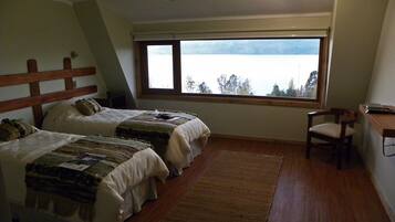 A bedroom with two beds, a wooden headboard, a desk, a chair, a large window with a view of a lake, and a ceiling fan.
