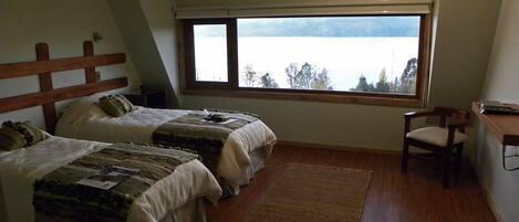 A bedroom with two beds, a wooden headboard, a desk, a chair, a large window with a view of a lake, and a ceiling fan.