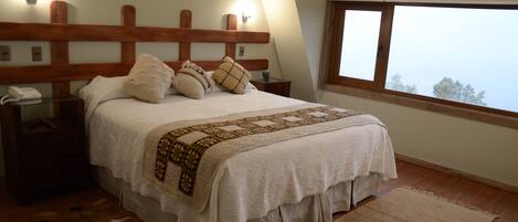 Un dormitorio con cabecera de madera, un ventanal grande, cama con ropa de cama blanca, una manta decorativa estampada y un tapete de cuero.