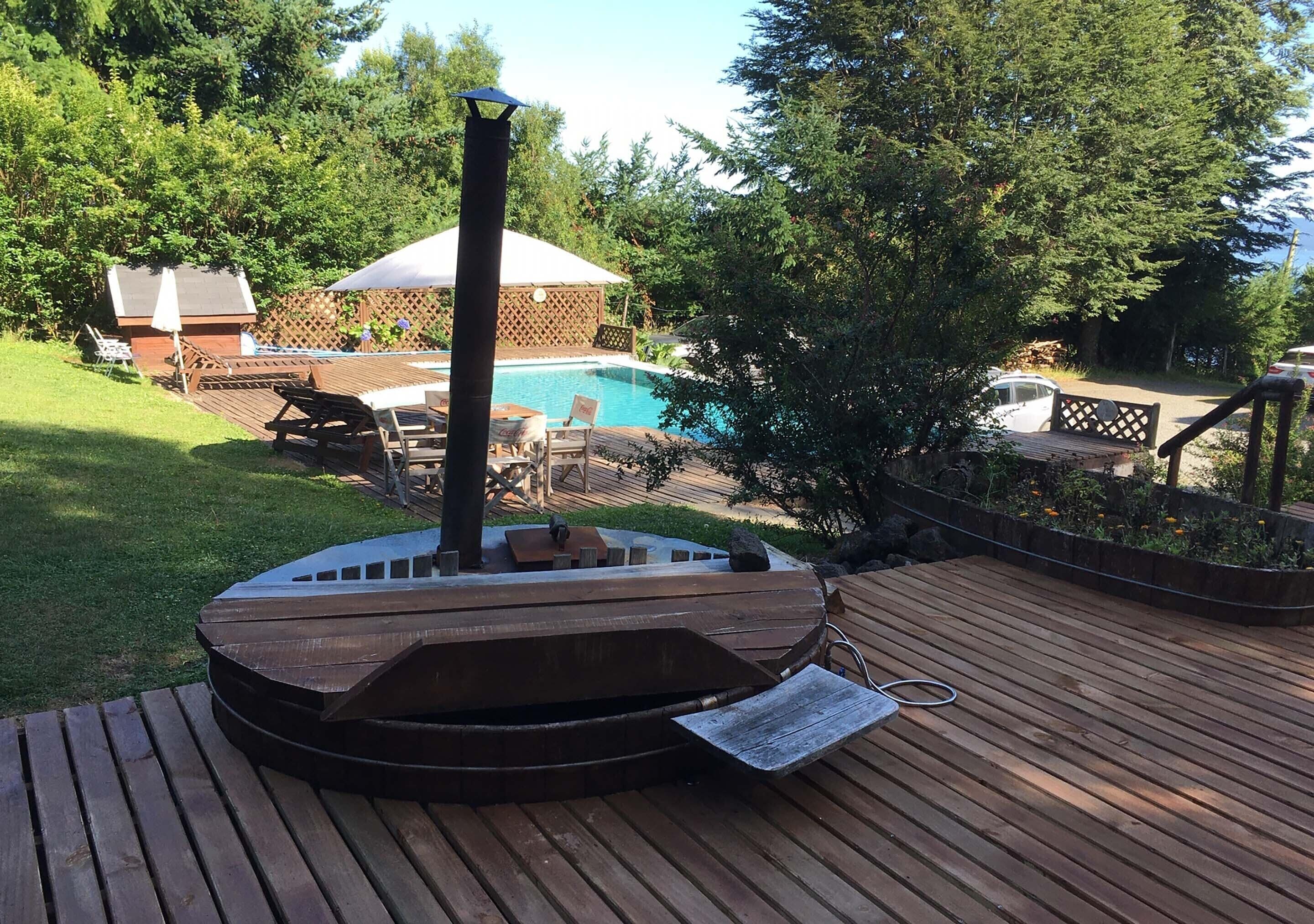 A hot tub on a wooden deck with a view of a backyard pool and trees.