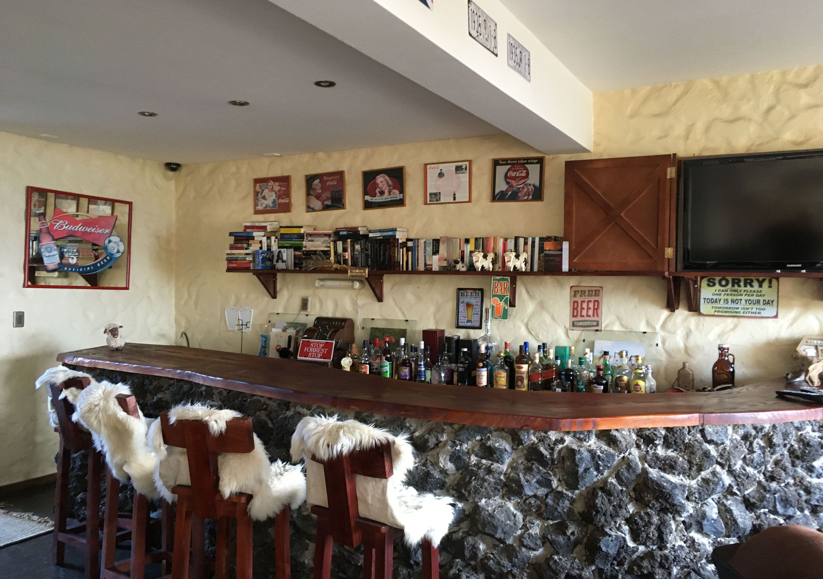A bar area with a stone counter, wooden bar stools, and a variety of bottles. There are framed pictures on the wall and a television mounted above the counter.