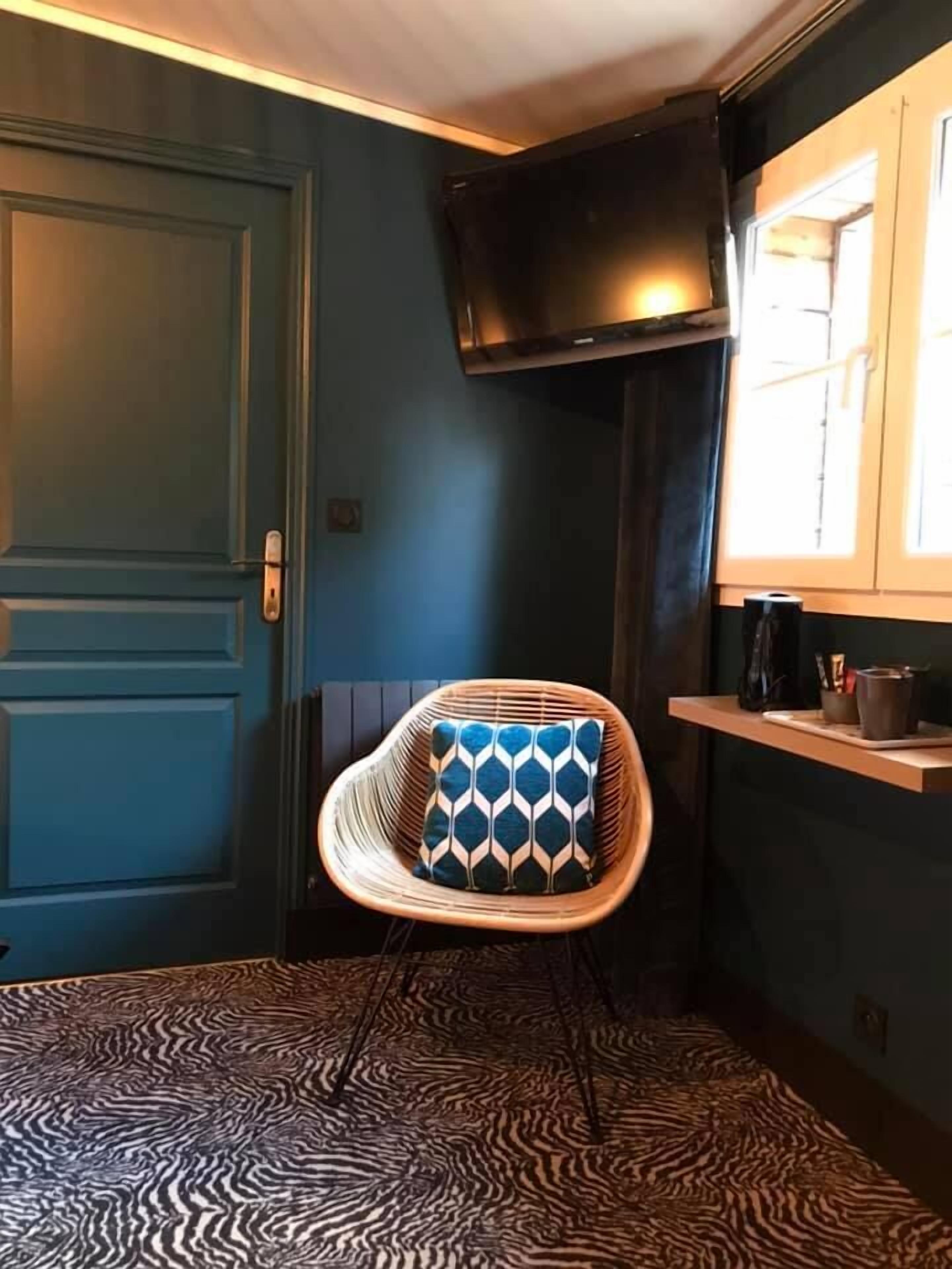Une pièce avec une porte bleue, une chaise en osier avec un coussin à motifs, une moquette à motifs, une étagère avec une bouilloire et un téléviseur fixé au mur.