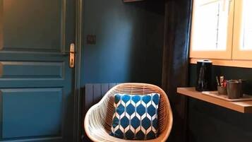 Une pièce avec une porte bleue, une chaise en osier avec un coussin à motifs, une moquette à motifs, une étagère avec une bouilloire et un téléviseur fixé au mur.