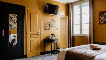 A hotel room with a bed, a television mounted on the wall, a small table with a kettle and bottles, and a window with curtains.