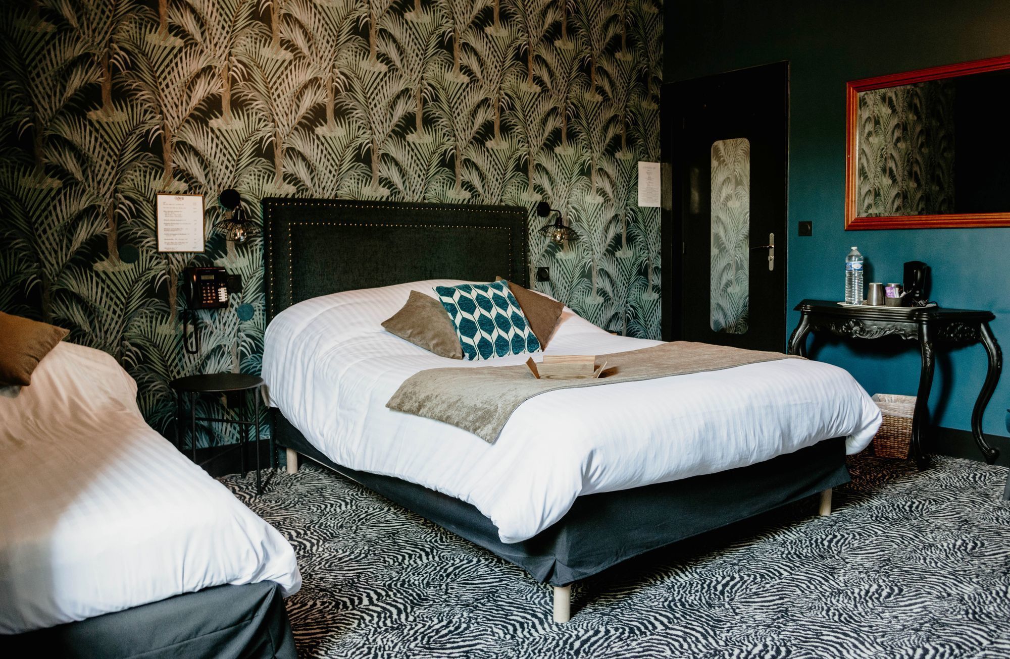 A hotel room with a large bed, a bedside table with a lamp, a telephone, and a wall with a tropical leaf pattern.