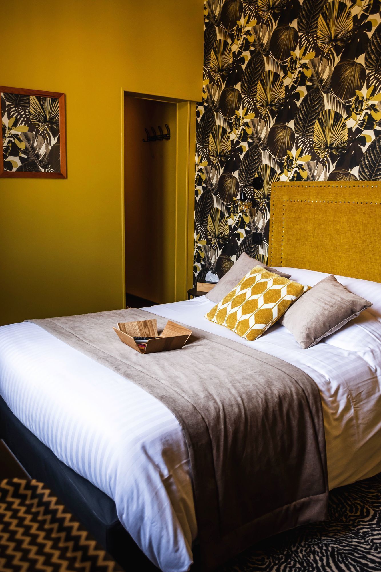 Habitación de hotel con cama, mesita de madera, cabecera y pared con estampado de hojas tropicales.
