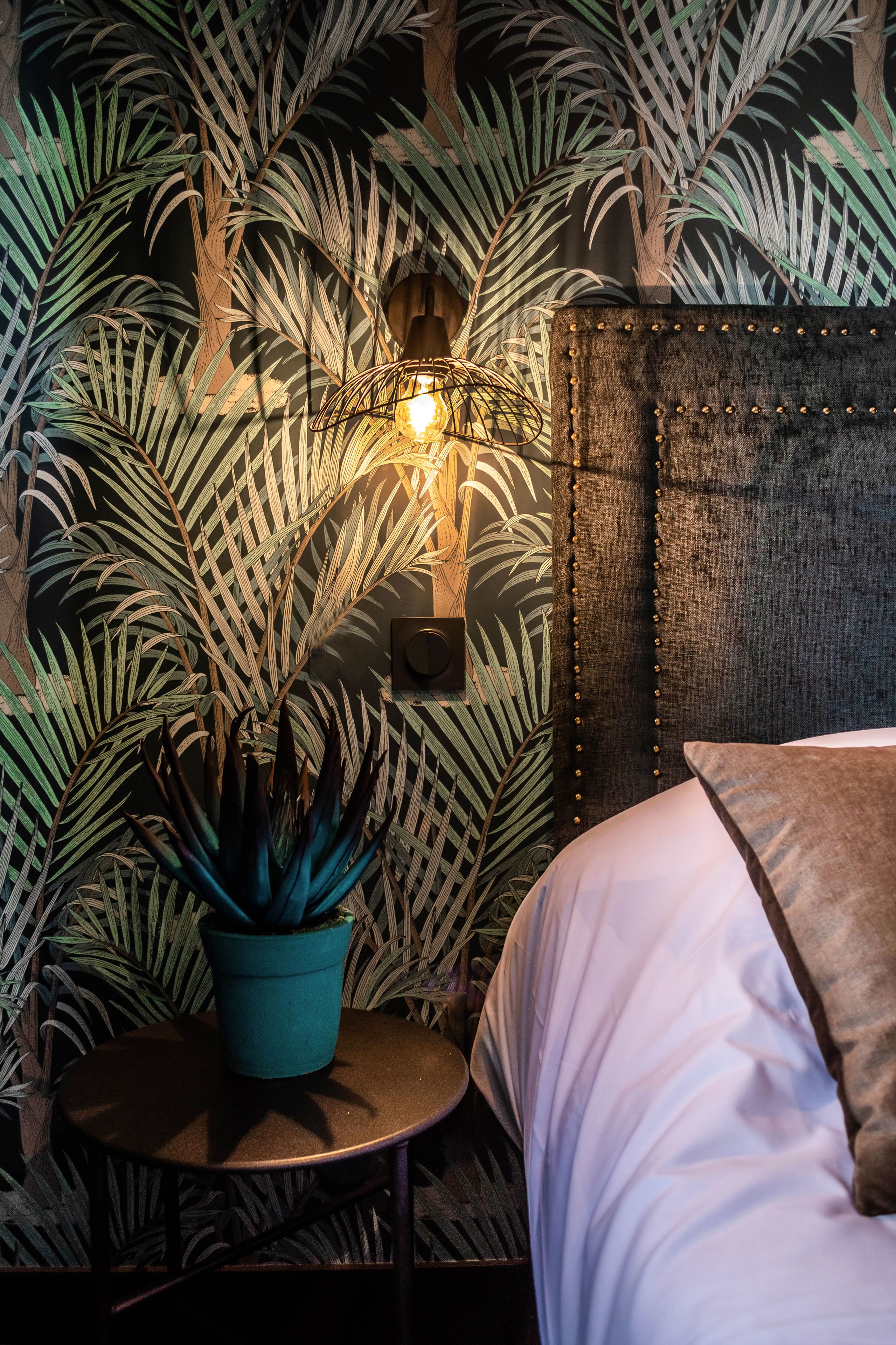 Une pièce comprenant un lit, une petite table ronde ornée d’un pot de plante, et un mur décoré d’un motif de feuilles tropicales.