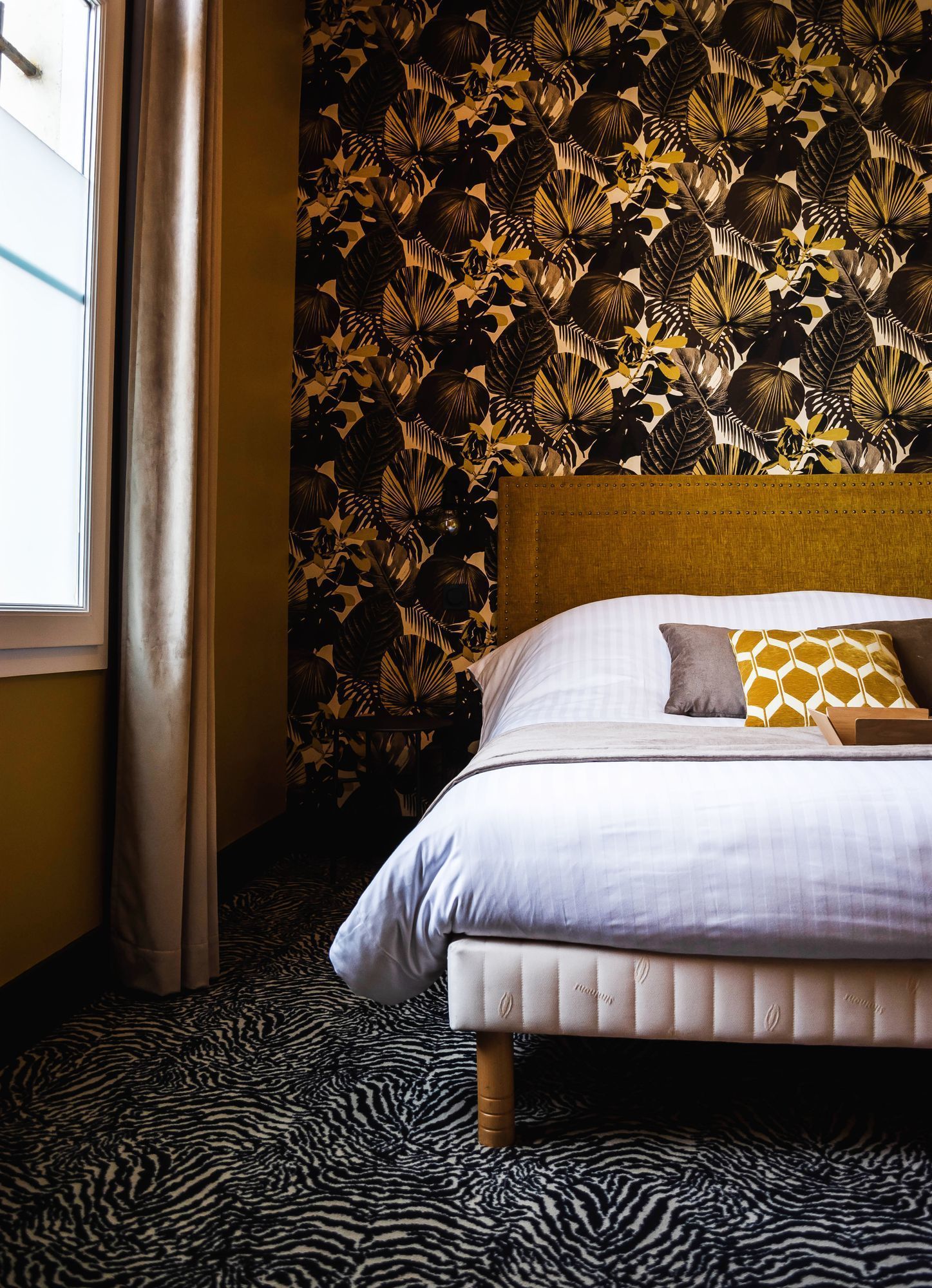 Ein Hotelzimmer mit einem zebra-gemusterten Teppich, einem Bett mit weißer Bettwäsche und gemusterten Kissen sowie einer Wand mit tropischem Blatt-Tapete.