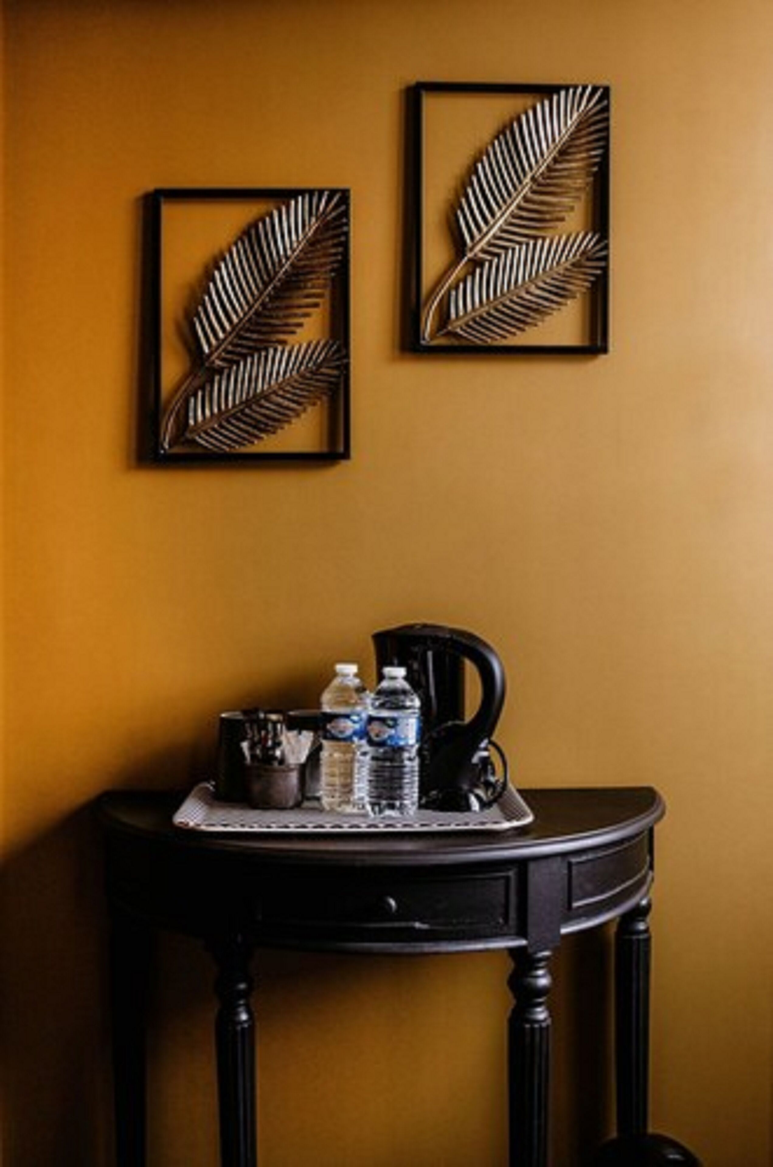 Une pièce avec une table en bois foncé adossée à un mur jaune moutarde, ornée de deux motifs de feuilles encadrés et d’un plateau contenant des bouteilles d’eau et une tasse.