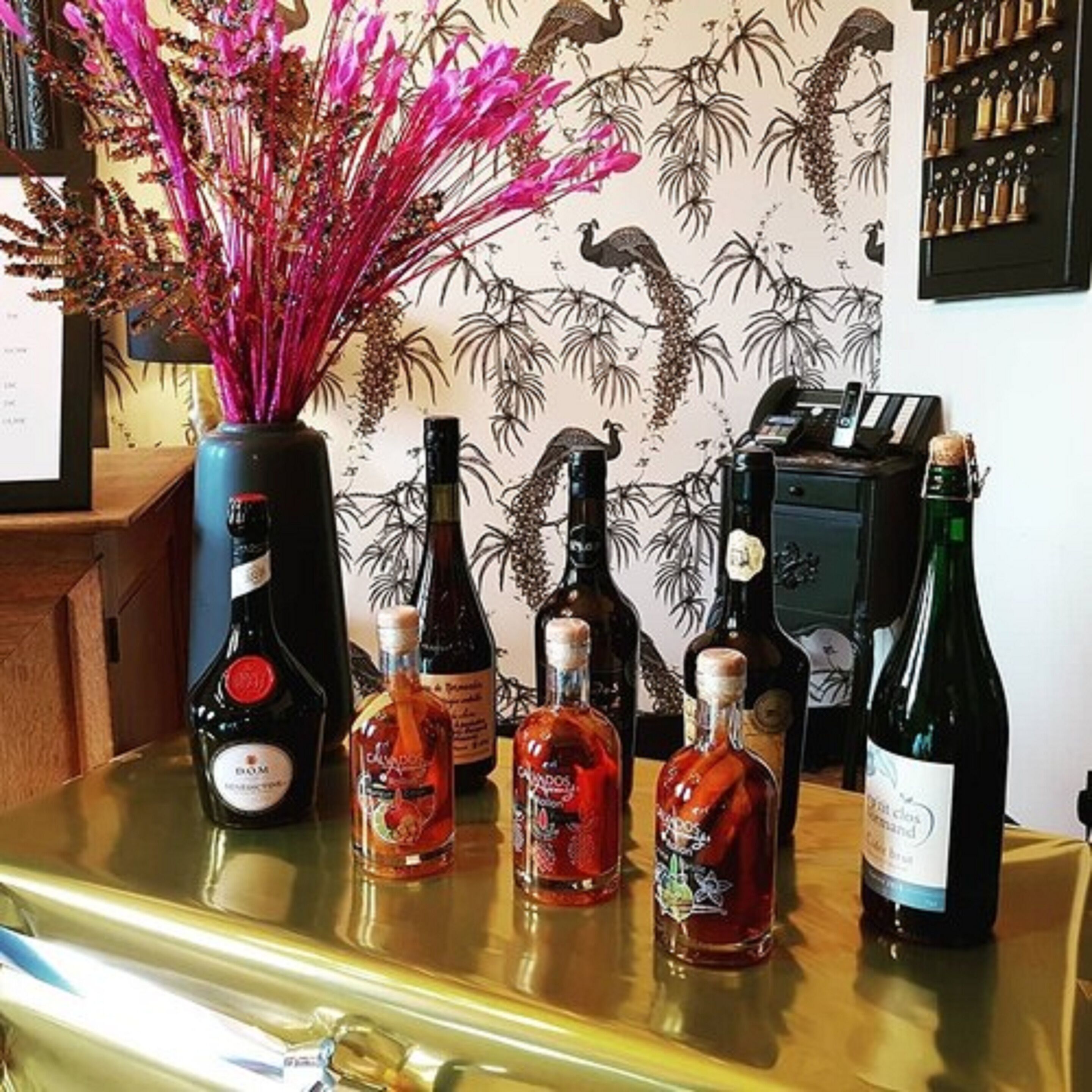 A bar area with bottles of liquor, a decorative vase with pink flowers, and a wall with a tropical wallpaper design.