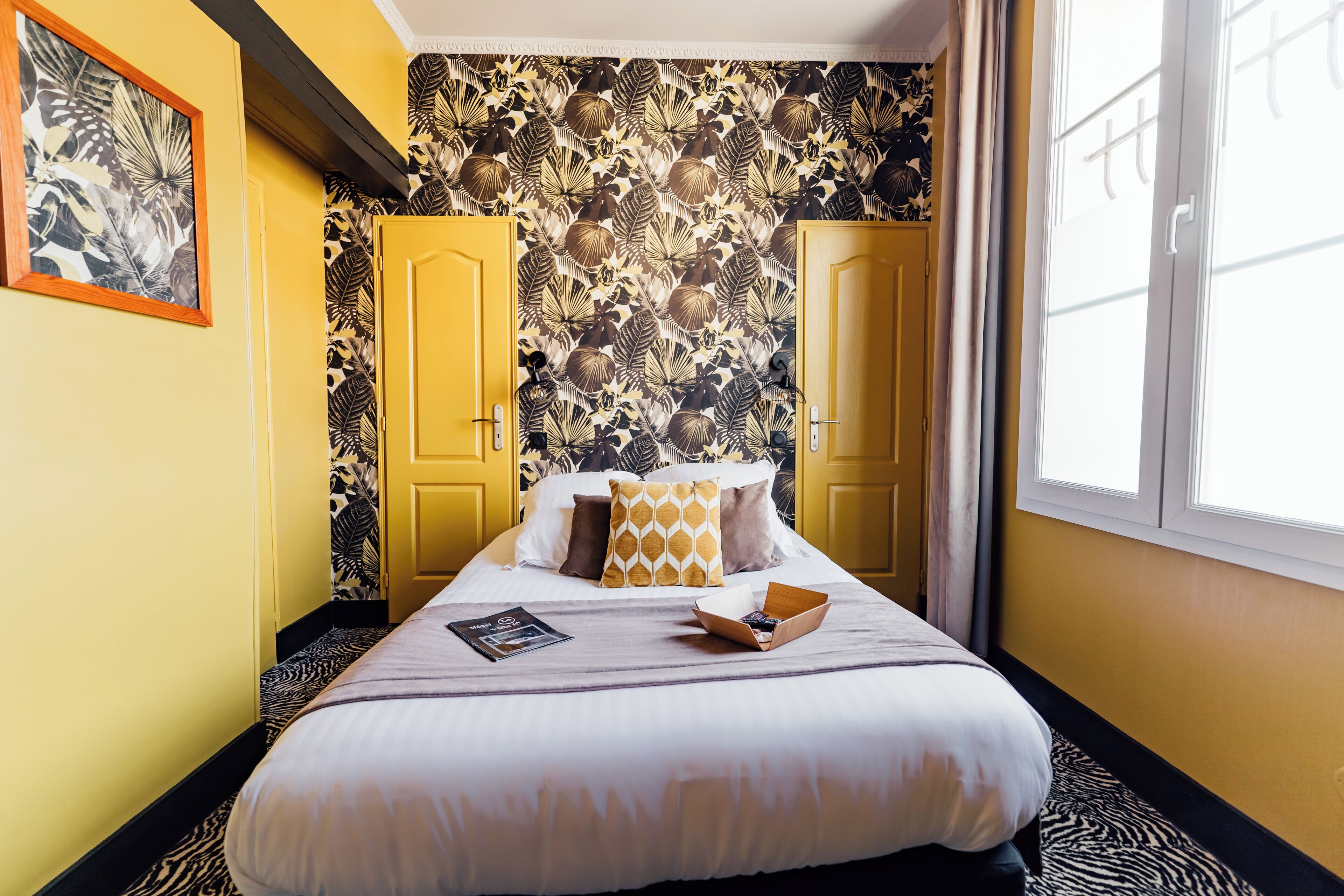 A hotel room with a bed, yellow doors, a patterned wallpaper, and a window.