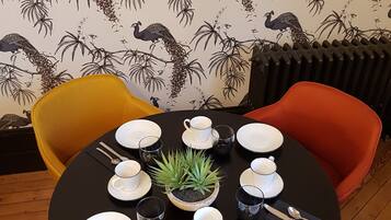 A round dining table with white plates, cups, and cutlery, surrounded by colorful chairs. A small potted plant is on the table.
