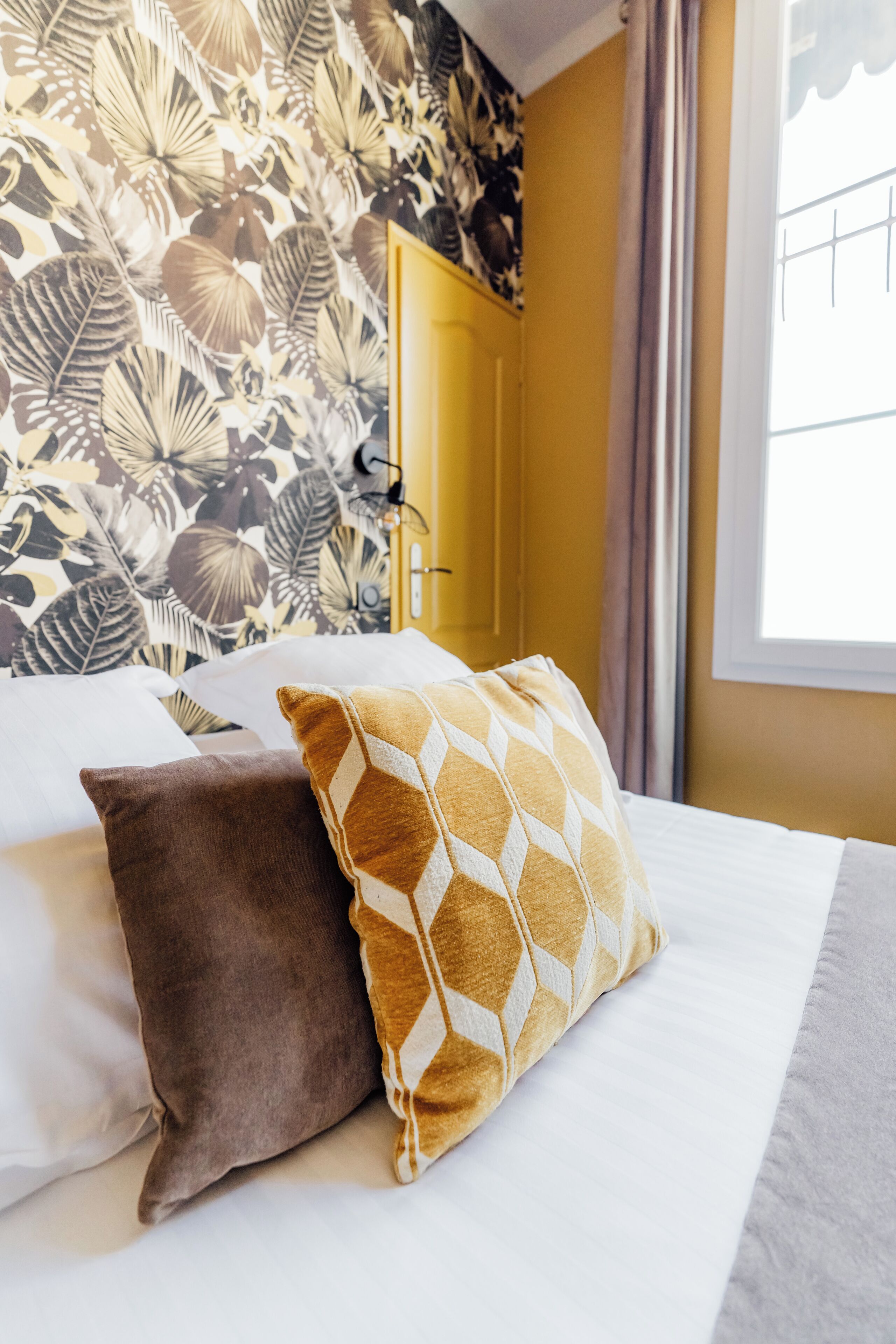 A bed with two pillows, a floral wallpaper, a yellow door, and a window with curtains.