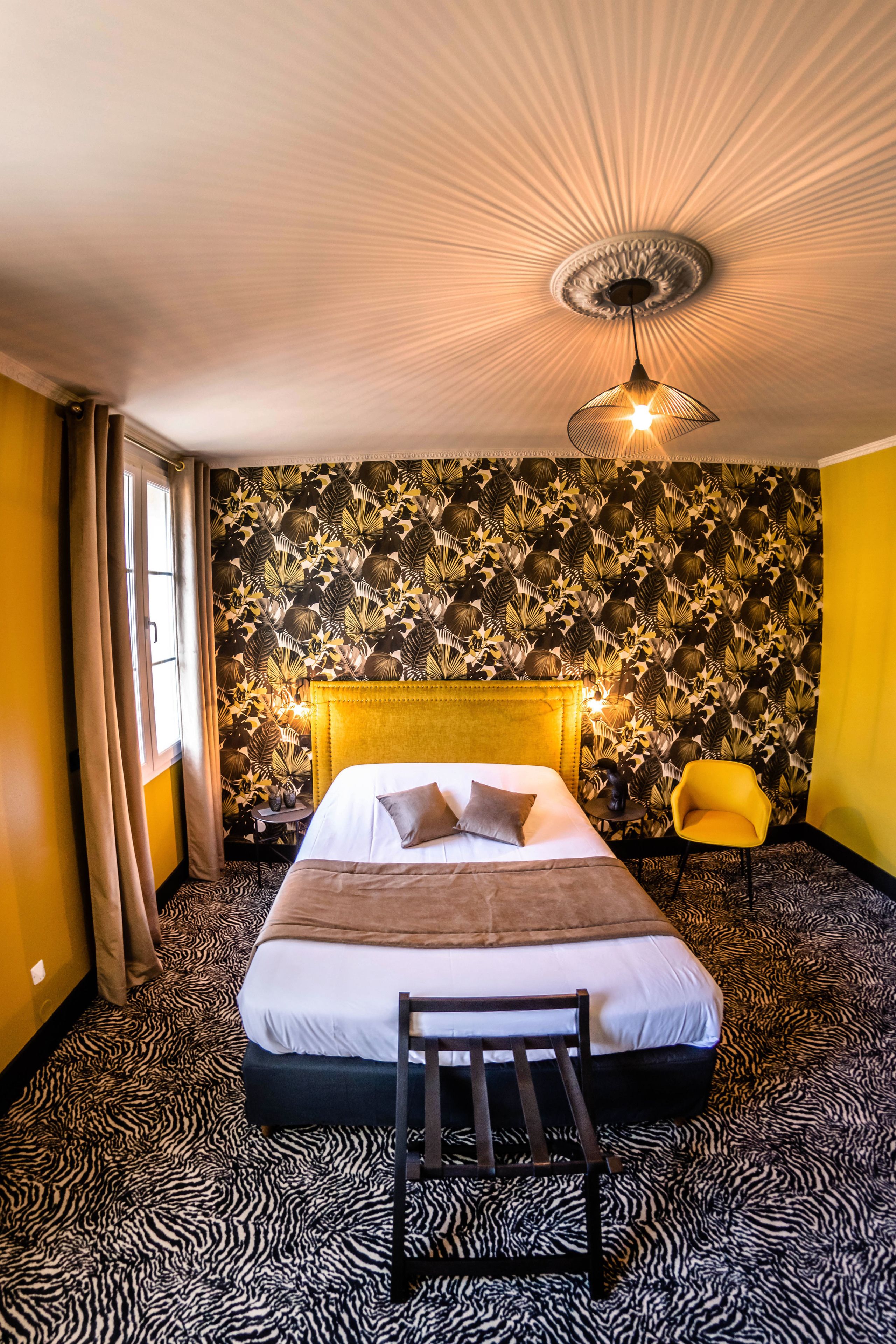 Una habitación de hotel con alfombra de estampado de cebra, una cama con cabecero, una silla amarilla y una lámpara en el techo.