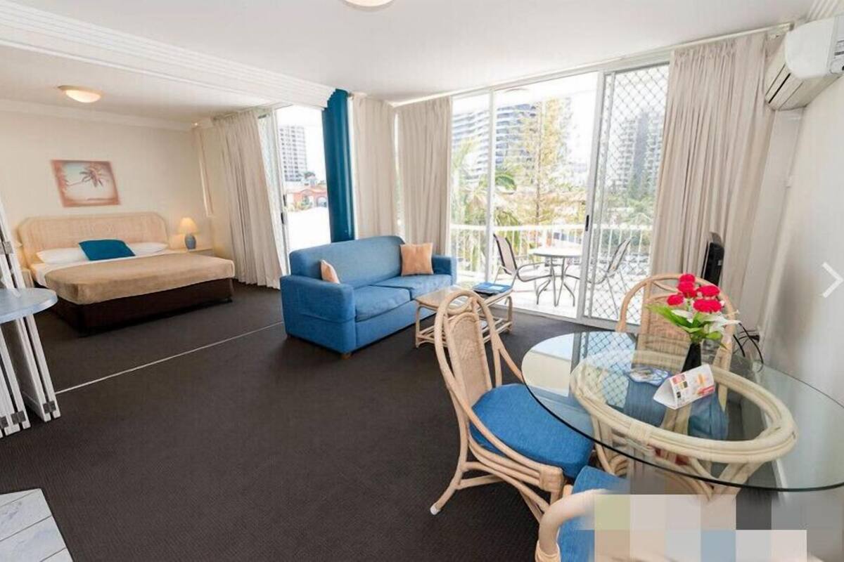 A modern hotel room with a bed, a blue sofa, a glass table, wicker chairs, and a balcony with a view of the city.
